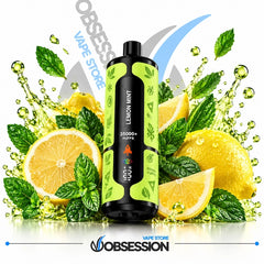 A disposable vape device labeled 'LEMON MINT' stands centered in front of a stylized fruit and mint splash scene with lemons, mint leaves, and water droplets. The vape has a black body with bright lime green side panels and a dark mouthpiece.