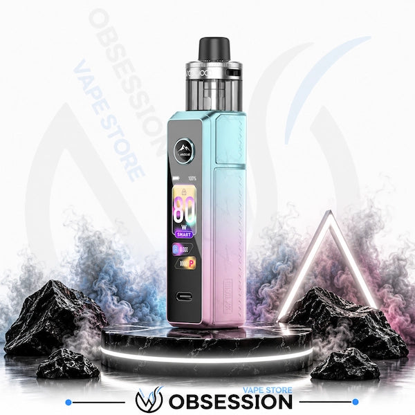 A Obsession vape device with a pastel blue and pink gradient body, standing on a black marble platform with digital smoke effects and rocks in the background.