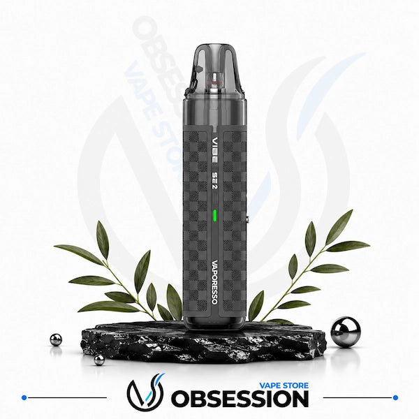 A dark gray Vibe 2 disposable vape device with a checkered pattern, displayed on a black marble base with decorative green leaves and metallic spheres against a white background with a faint store watermark.