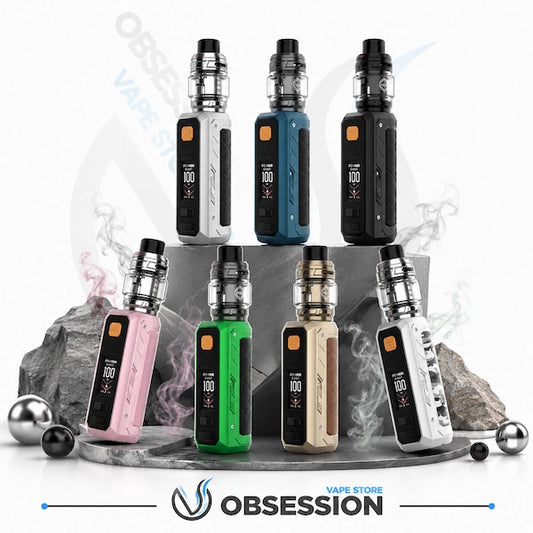 Six vape devices in different colorways are arranged on a gray pedestal with rocks and metallic balls against a white background. The devices include black, silver, blue, pink, green, and beige models, each with a digital display and a clear tank.